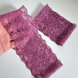 May include: Close-up of two pieces of mauve lace fabric. The lace has a floral pattern and a scalloped edge. The fabric is held up against a white background.
