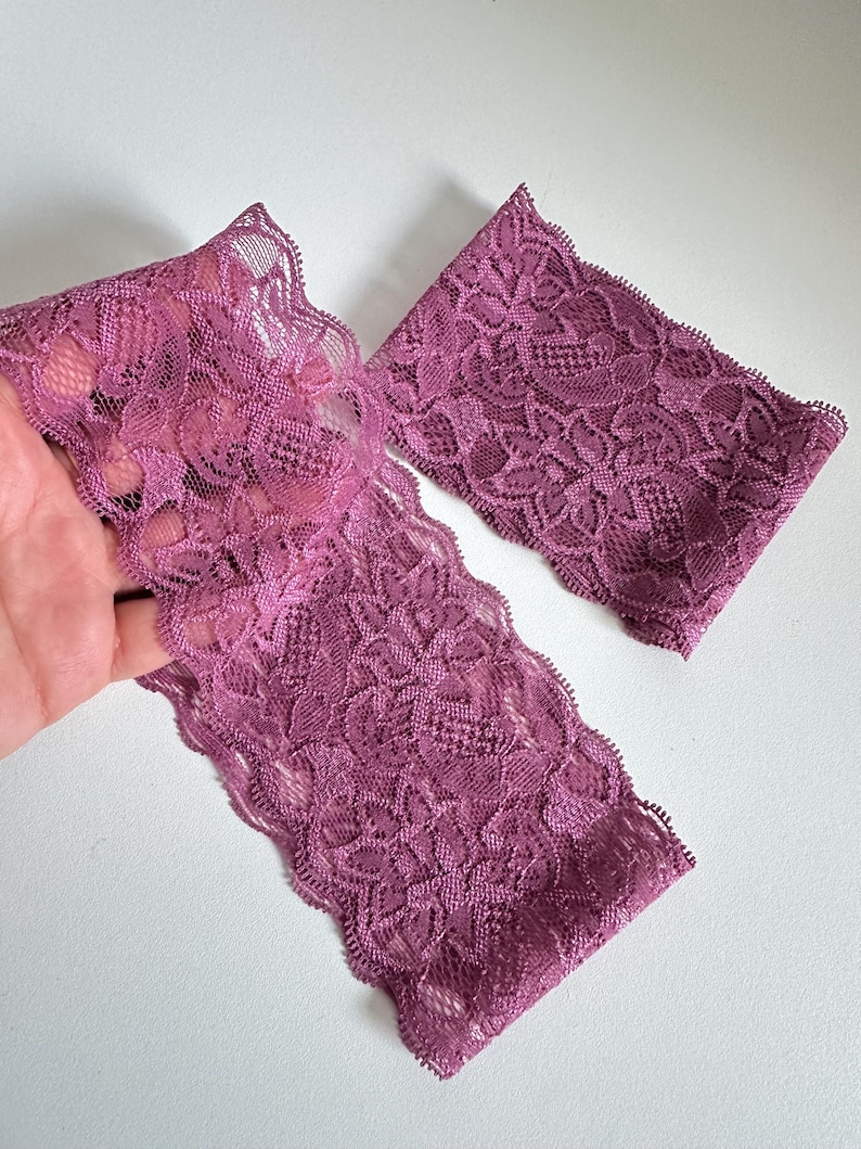 May include: Close-up of two pieces of mauve lace fabric. The lace has a floral pattern and a scalloped edge. The fabric is held up against a white background.
