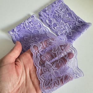 May include: Three pieces of lavender lace fabric with a floral pattern. The delicate lace is held in a hand, showcasing its intricate design and soft texture. The fabric is a light purple color.
