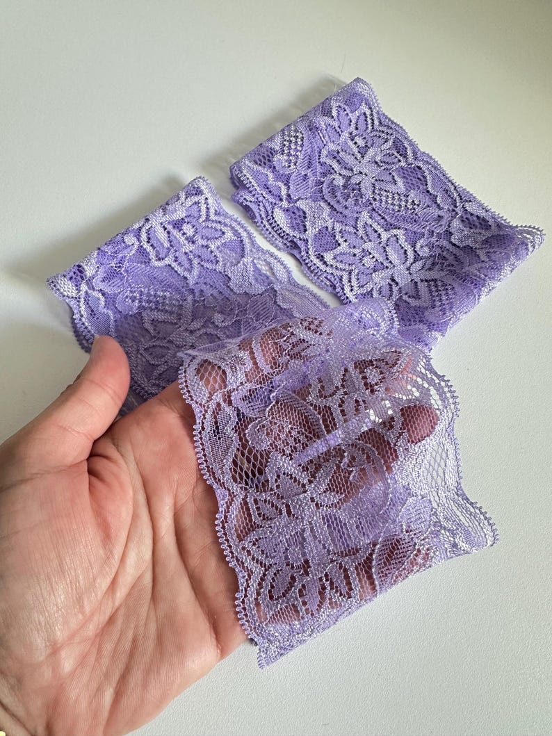 May include: Three pieces of lavender lace fabric with a floral pattern. The delicate lace is held in a hand, showcasing its intricate design and soft texture. The fabric is a light purple color.