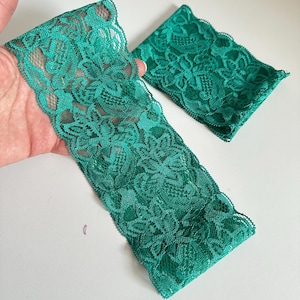 May include: Two pieces of emerald green lace fabric. One piece is a long strip, and the other is a smaller square. The lace has a floral pattern and a scalloped edge. The fabric is held in a hand against a white background.