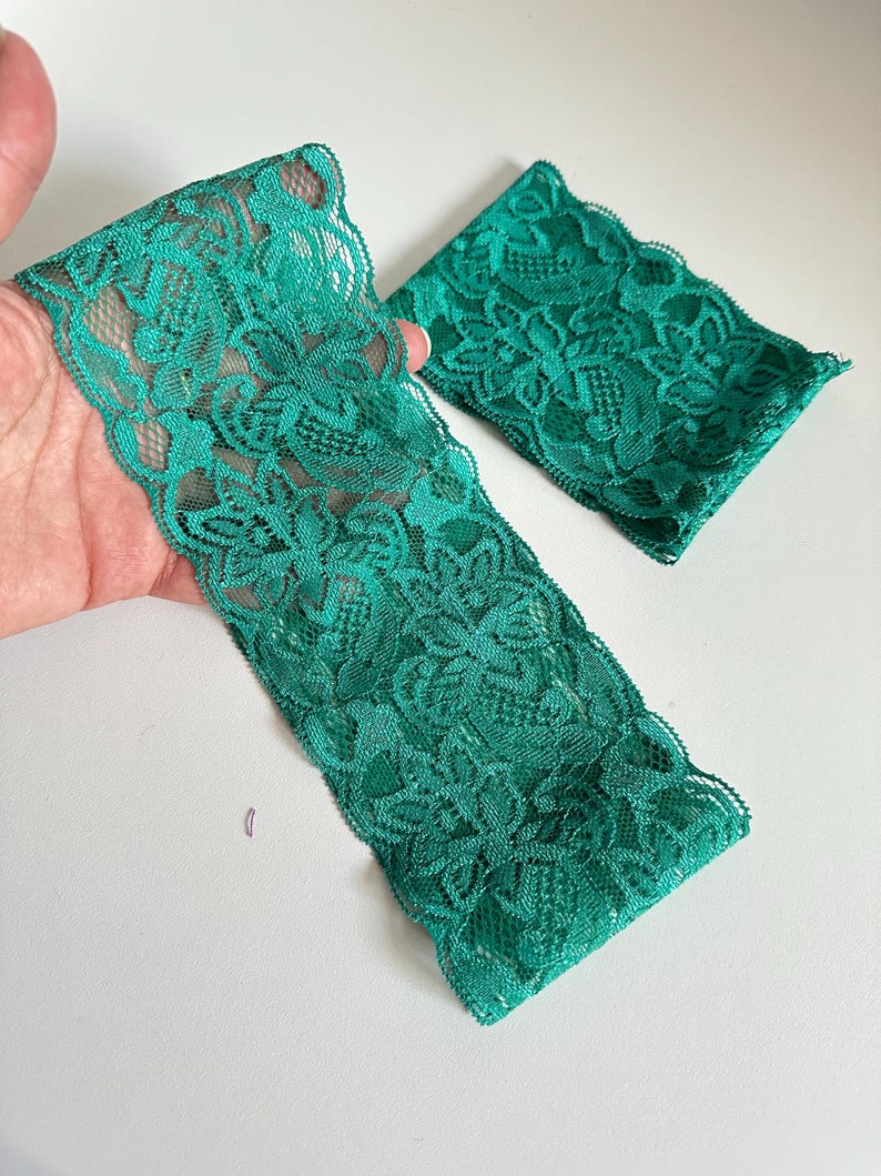May include: Two pieces of emerald green lace fabric. One piece is a long strip, and the other is a smaller square. The lace has a floral pattern and a scalloped edge. The fabric is held in a hand against a white background.