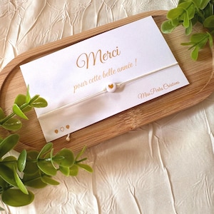 Könnte beinhalten: Ein weißes Schnurarmband mit einer einzelnen runden weißen Perle mit goldenem Herz, befestigt an einer weißen Karte mit dem Text Merci pour cette belle année und Miss Perles Créations, verziert mit kleinen goldenen Herzsymbolen unten links.