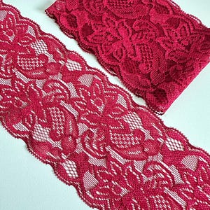 May include: Two pieces of crimson red lace fabric. The lace features a floral pattern with a scalloped edge. The fabric is displayed on a white surface, showcasing the intricate design and texture of the lace.