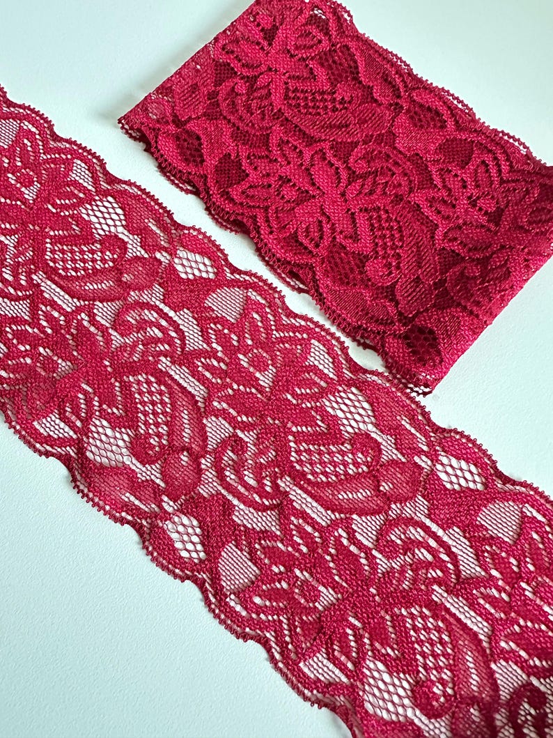 May include: Two pieces of crimson red lace fabric. The lace features a floral pattern with a scalloped edge. The fabric is displayed on a white surface, showcasing the intricate design and texture of the lace.