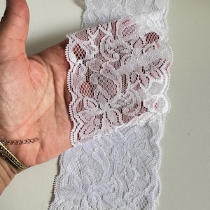 May include: White lace trim with a floral pattern. The lace is held in a hand, showcasing the delicate design and scalloped edges. This trim is suitable for various sewing and craft projects.