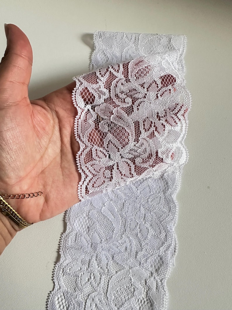 May include: White lace trim with a floral pattern. The lace is held in a hand, showcasing the delicate design and scalloped edges. This trim is suitable for various sewing and craft projects.