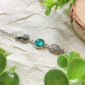 Può includere: Un bracciale argentato con una pietra centrale ovale color turchese, affiancata da due ciondoli a forma di ali dettagliate. Il bracciale è posto su uno sfondo crema testurizzato, con fogliame verde e una fetta di legno sullo sfondo.