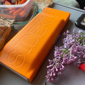 Könnte beinhalten: Ein orangefarbenes rechteckiges Etui mit dem Wort "SEK" auf der Oberseite. Das Etui liegt auf einer grauen Oberfläche, neben einem Behälter mit Karottensticks und einem Zweig lila Flieder.