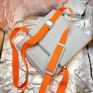 May include: A gray rectangular bag with bright orange straps and silver metal buckles. The straps are adjustable and designed to be worn over the shoulders. The bag is set against a crinkled silver background.