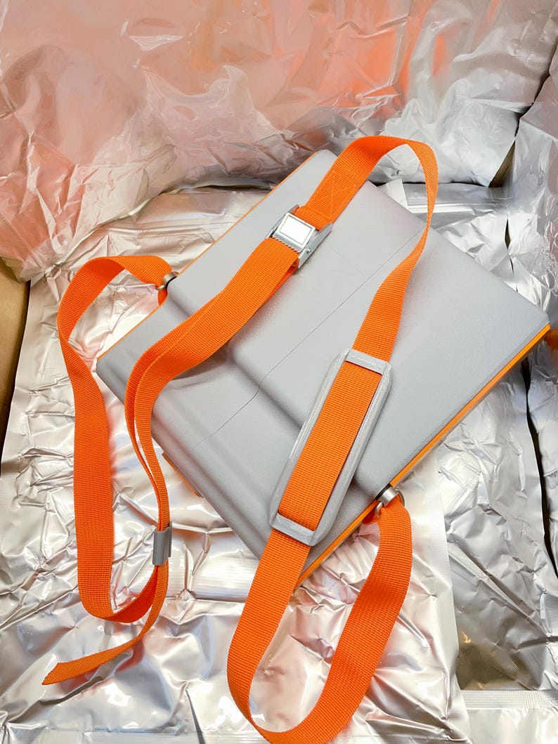 May include: A gray rectangular bag with bright orange straps and silver metal buckles. The straps are adjustable and designed to be worn over the shoulders. The bag is set against a crinkled silver background.