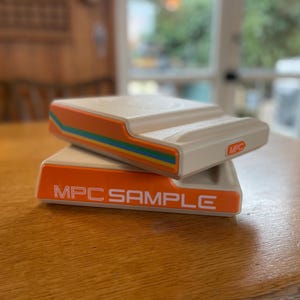 Peut inclure: Deux appareils électroniques beiges empilés sur une table en bois. L'appareil supérieur a une bande orange avec des accents bleus et verts. L'appareil inférieur porte l'inscription "MPC SAMPLE" en blanc sur fond orange. Les appareils portent l'inscription "MPC".