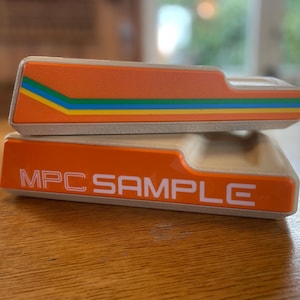 May include: Two orange MPC SAMPLE electronic music production devices are stacked on a wooden surface. The top device has a colorful stripe design. The bottom device has the text "MPC SAMPLE" in white.