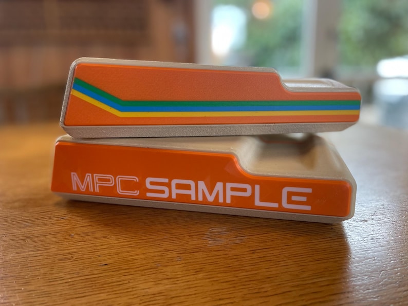 May include: Two orange MPC SAMPLE electronic music production devices are stacked on a wooden surface. The top device has a colorful stripe design. The bottom device has the text "MPC SAMPLE" in white.