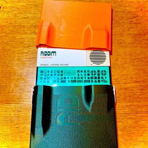 May include: An orange plastic case with the word "RIDDIM" and "SUPERTONE" printed on a white card. Below the white card is a teal card with symbols and text. A black case is at the bottom.