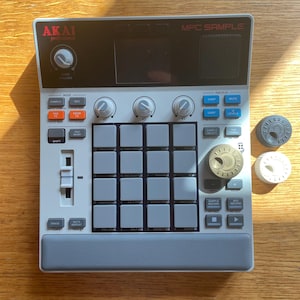 May include: A gray and white Akai MPC Sample music production device with numerous buttons, knobs, and a display screen. Three circular knobs are next to the device. The device has the text "MPC SAMPLE" and "AKAI professional".