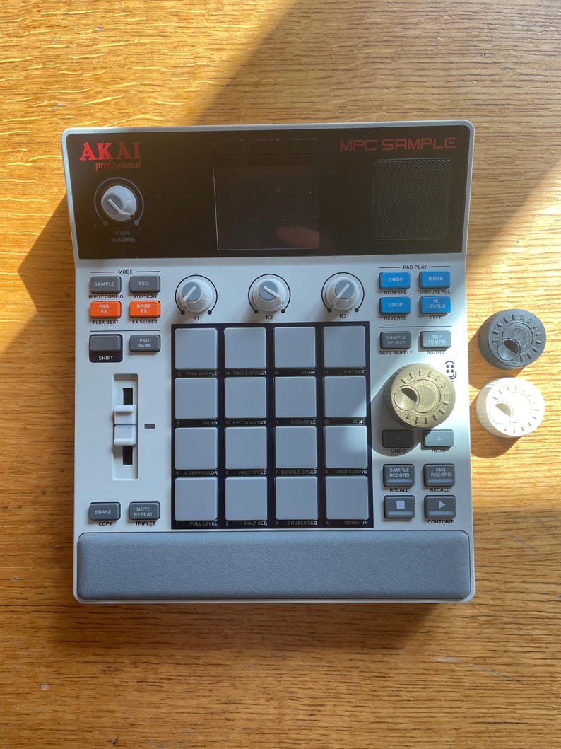 May include: A gray and white Akai MPC Sample music production device with numerous buttons, knobs, and a display screen. Three circular knobs are next to the device. The device has the text "MPC SAMPLE" and "AKAI professional".