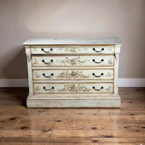 May include: A vintage, off-white painted chest of drawers with four drawers. Each drawer is adorned with hand-painted floral and musical instrument motifs. The chest has black, curved handles and sits on a wooden floor.