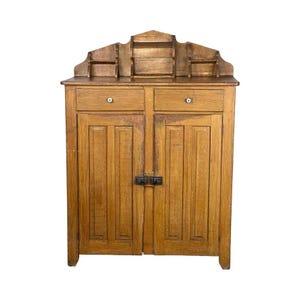 May include: A vintage wooden cabinet with two drawers and two doors. The cabinet is made of light brown wood and has a decorative top with small shelves. The doors are secured with a black latch.