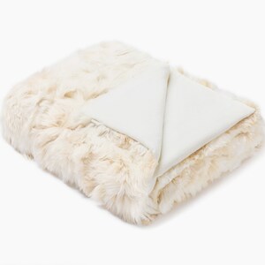 May include: A white faux fur throw blanket with a soft, plush texture. The blanket is folded in half and has a white underside.