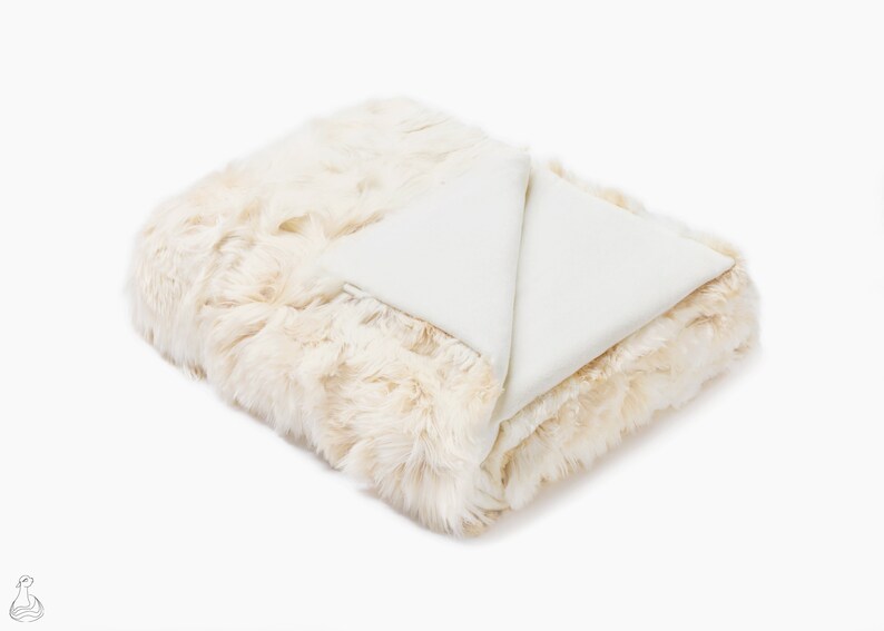May include: A white faux fur throw blanket with a soft, plush texture. The blanket is folded in half and has a white underside.