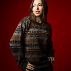Op de afbeelding: Een vrouw draagt een bruine, blauwe en zwarte trui met een ronde hals. De trui heeft een fair isle-patroon.
