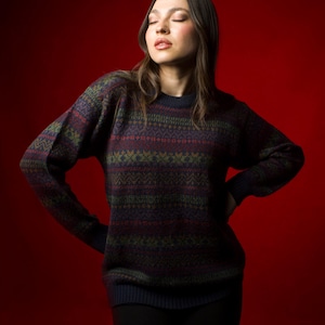 Könnte beinhalten: Ein gemusterter Pullover in Marineblau, Burgunderrot, Grün und Gold. Der langärmlige Pullover hat einen Rundhalsausschnitt und ein geometrisches Design. Der Hintergrund ist einfarbig rot.