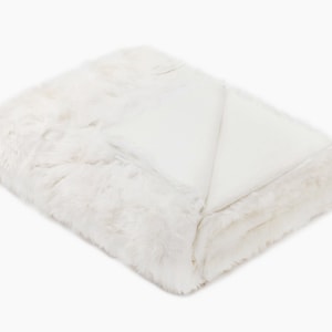 May include: A white faux fur throw blanket with a soft, fluffy texture. The blanket is folded in half and has a smooth, white underside.