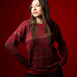 May include: A woman with long brown hair is wearing a red and green patterned sweater with long sleeves. She is standing in front of a red background.