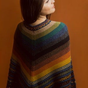 Pu&ograve; includere: Un poncho a righe multicolore con frange. Il poncho, con scollo a V, presenta sfumature di blu, verde, arancione e giallo. Ha un bordo fantasia ed &egrave; indossato da una persona, su uno sfondo arancione.