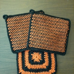 Puede incluir: Dos agarraderas de olla de ganchillo en negro y naranja. Una agarradera tiene un diseño cuadrado y la otra tiene un diseño cuadrado con un cuadrado más pequeño en el centro.