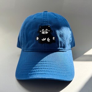 Könnte beinhalten: Blaue Baseballkappe mit einem aufgestickten schwarzen Kätzchen auf der Vorderseite. Das Kätzchen hat weiße Akzente und sitzt. Die Kappe hat einen gebogenen Schirm und einen verstellbaren Riemen.