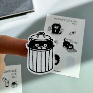 Könnte beinhalten: Ein schwarz-weißer Aufkleber mit einer Katze, die aus einem Mülleimer schaut. Der Aufkleber ist Teil der "Meowffice Life"-Serie, mit weiteren Aufklebern im Hintergrund. Die Katze hat große Augen und ist im Cartoon-Stil gezeichnet.