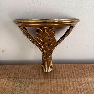 Puede incluir: Un estante de pared de color dorado con una superficie superior semicircular sostenida por un soporte decorativo en forma de manojo de trigo. La estructura tiene un acabado metálico brillante e incluye tallas detalladas en forma de hojas.