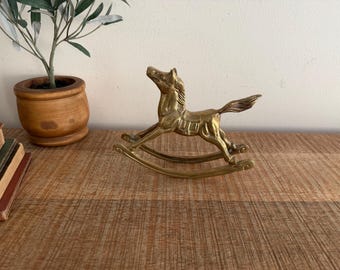 Puede incluir: Una figura de caballo balancín de latón con una melena y cola detalladas. El caballo está montado sobre balancines curvos, y el diseño general es vintage. La figura se exhibe sobre una superficie de madera, con una planta en maceta y libros en el fondo.