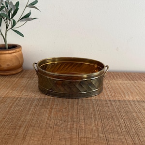 Könnte beinhalten: Ovale Messing-Pflanzschale mit strukturierter, rautenförmiger Außenseite und zwei kleinen Griffen. Der Innenraum ist goldfarben glänzend. Ein kleiner Holztopf mit einer Pflanze befindet sich im Hintergrund.