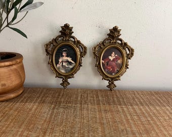 May include: Two ornate gold-toned picture frames with intricate details. Each frame holds a portrait of a person in period clothing. The frames are hanging on a light brown wooden surface.