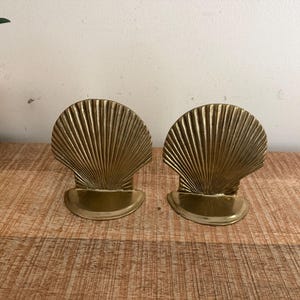 May include: A pair of polished brass bookends shaped like open scallop shells. Each bookend features a semi-circular base and a vertical, fan-shaped shell design with radiating ridges, showcasing a metallic gold finish and a smooth, reflective surface.