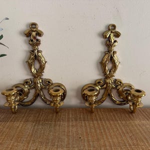 May include: Two ornate, gold-colored wall sconces. Each sconce features a decorative top, a central design, and two candle holders. The sconces are made of metal and are mounted on a light-colored wall. The sconces are displayed on a wooden surface.