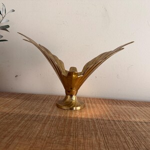 Puede incluir: Una escultura de latón pulido de un ave con las alas completamente extendidas en posición de vuelo ascendente. El ave está montada sobre una base circular de pedestal, con un acabado metálico dorado liso y una textura visible.