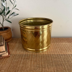 May include: A cylindrical brass planter featuring a polished finish and a decorative diagonal ribbed texture around the exterior. The metal container has a wide open top and a smooth, reinforced rim, showcasing a bright, reflective gold-toned surface.