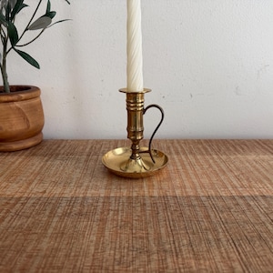 May include: A vintage brass candlestick holder with a curved handle and a round base. A white, twisted taper candle is inserted in the holder. A small potted plant sits to the left of the candlestick on a wooden surface.