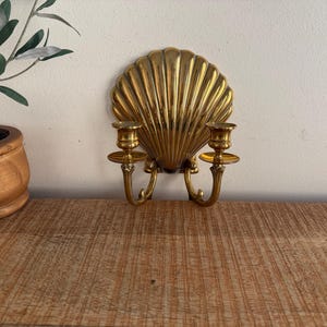 May include: A gold-colored wall sconce shaped like a seashell, with two candle holders. The sconce is mounted on a white wall, and a small potted plant is visible on a wooden surface.