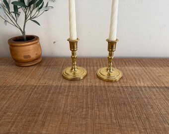 Può includere: Due candelabri in ottone con candele bianche a spirale. I candelabri hanno una base rotonda e uno stelo dettagliato. Un piccolo vaso di legno con una pianta è sullo sfondo. Gli oggetti sono su una superficie di legno.
