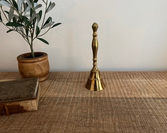 Pode incluir: Um sino de latão dourado com topo arredondado e base alargada. O sino está sobre uma superfície de madeira.