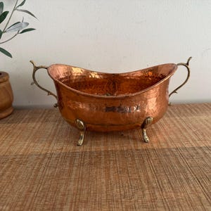 May include: A hammered copper bowl with a polished finish, featuring two ornate brass handles on the sides and four small brass feet shaped like shells. The oval-shaped vessel has a wide, open top and a deep, rounded basin for holding items.