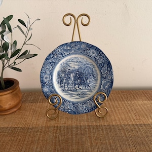 May include: A round ceramic plate featuring a blue and white transferware pattern of figures on horseback in a winter landscape, surrounded by a floral border, displayed on a gold-toned metal wire stand with decorative scrolled feet.