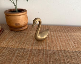 Può includere: Una statuetta a forma di cigno in ottone dorato con collo curvo e becco dettagliato. Il cigno è posizionato su una superficie di legno, con un vaso di legno contenente una pianta sullo sfondo. È un oggetto decorativo.