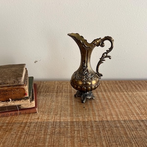 Puede incluir: Un pequeño cántaro de metal dorado con un diseño ornamentado. El cántaro tiene un asa y un pico. Está sentado sobre una superficie de madera.