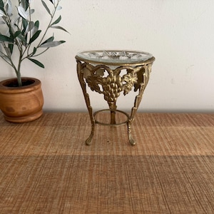 May include: A brass stand featuring a detailed relief of grape clusters and leaves, supporting a clear, circular glass bowl. The metal base has three curved legs connected by a circular ring, providing a stable structure for the glass insert.